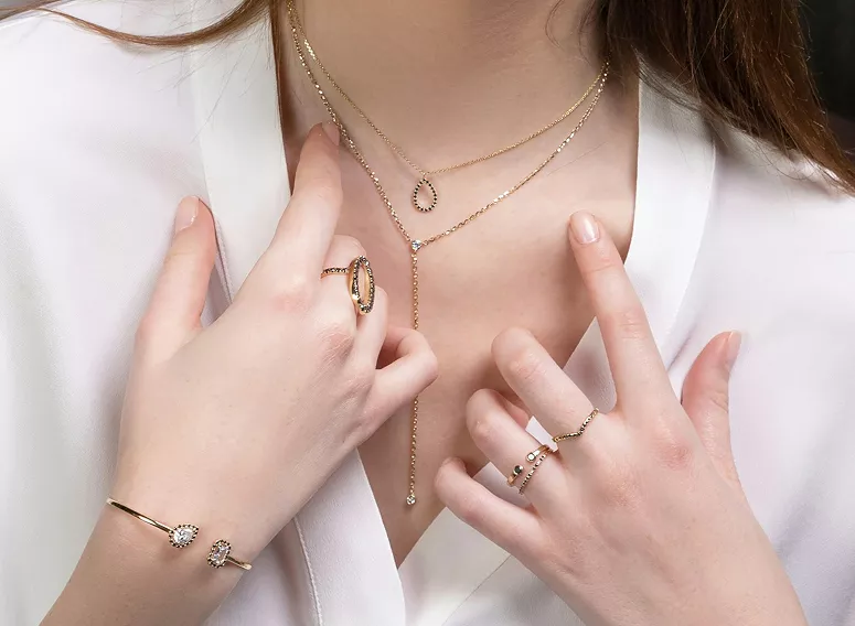 Woman in white shirt wearing layered gold necklaces with horseshoe pendant, delicate rings, and crystal-embellished bracelet jewelry.