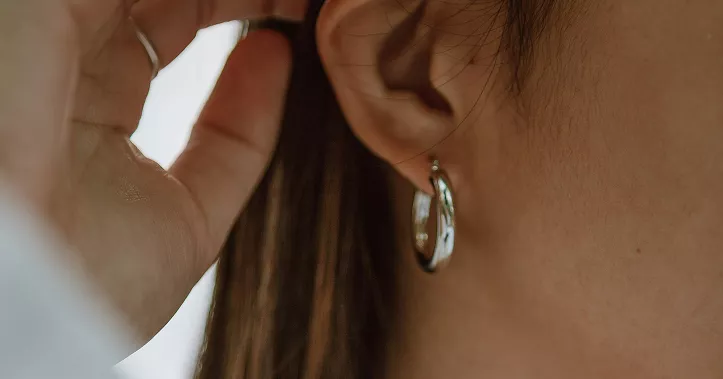 Side profile showing woman's ear with gold hoop earring featuring pearl accents, brown hair tucked behind ear in intimate beauty detail shot