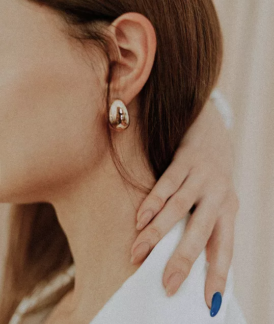 Close-up of woman wearing elegant gold dome earring with brown hair, showing neck and shoulder in minimalist white clothing style.