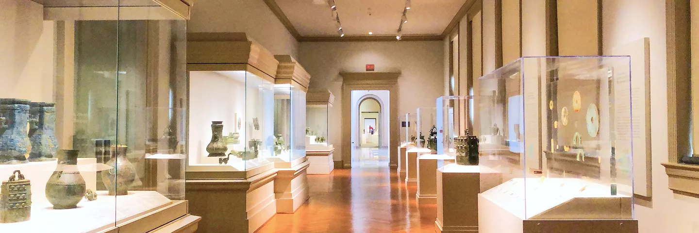 Museum gallery interior with illuminated display cases showcasing ancient pottery and artifacts, warm lighting, polished floors, columns.