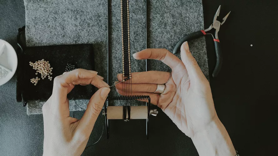 Jeweler's hands crafting necklace with precision tools, beads, and pliers on gray work surface, showcasing artisan jewelry making process.