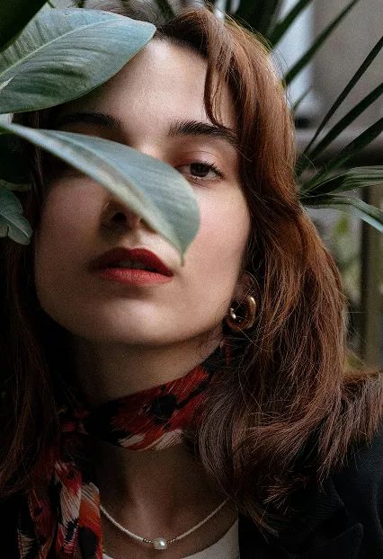 Woman with brown hair partially obscured by tropical leaves, wearing red lipstick, patterned scarf, and delicate necklace in natural portrait