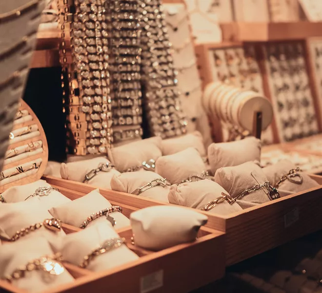 Jewelry store display with bracelets on beige cushioned stands, necklaces hanging in background, warm lighting in boutique setting.
