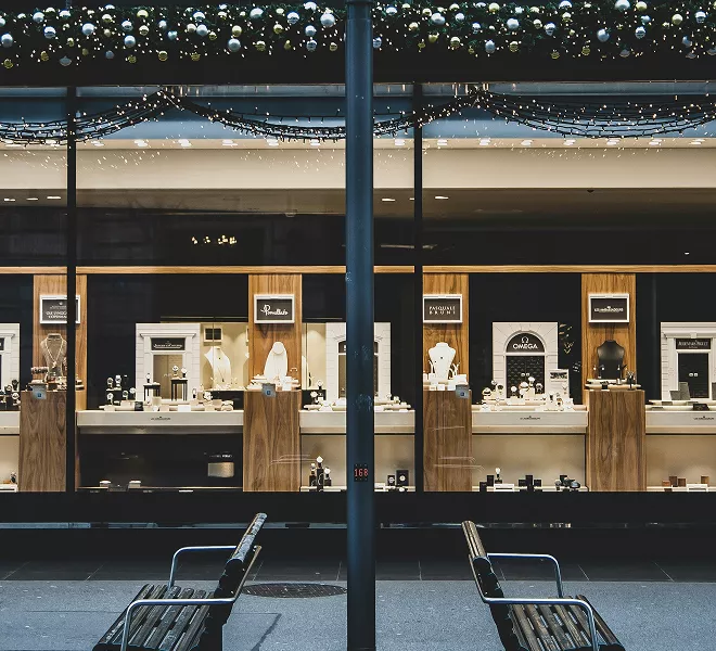 Jewelry store display windows with illuminated wooden showcases featuring luxury watches and accessories, viewed from outside mall