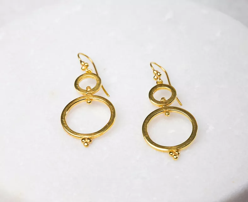 Pair of gold dangle earrings with double circle design and decorative granulation details on white marble surface in product photography