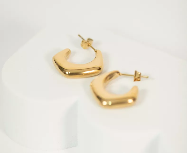 Polished gold chunky hoop earrings with curved geometric shape and post backs displayed on white marble in minimalist jewelry photography