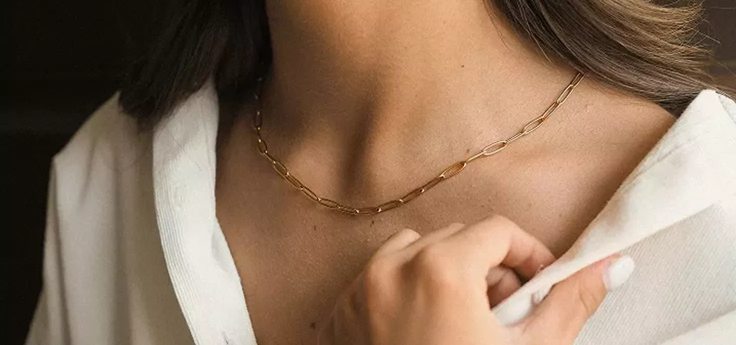 Close-up of woman wearing delicate gold chain necklace with white ribbed sweater, hand gently touching collar in elegant lifestyle portrait