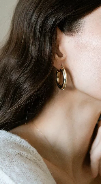Side profile showing woman's ear with gold hoop earring featuring pearl accents, brown hair tucked behind ear in intimate beauty detail shot