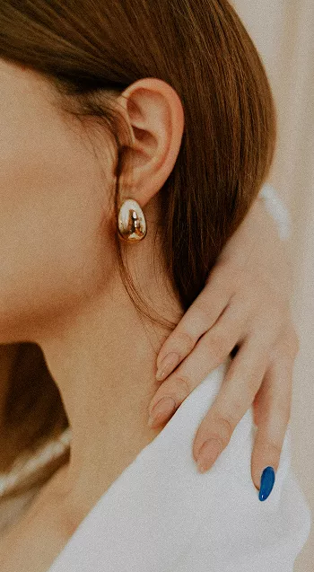 Side profile showing woman's ear with gold hoop earring featuring pearl accents, brown hair tucked behind ear in intimate beauty detail shot
